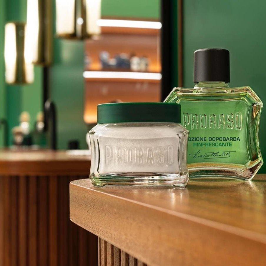 En flaska Proraso After Shave Lotion - Uppfriskande (grön) och en burk tvål sitter på ett träbord bredvid lite rakvatten.